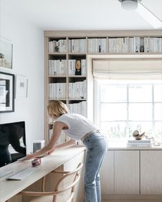 a woman leaning over a desk in front of a computer and bookshelf filled with books