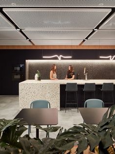 two women sitting at a bar in an office setting with chairs and plants on the floor