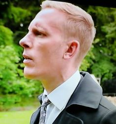 a man with blonde hair wearing a suit and tie in front of some trees looking off into the distance