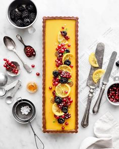 a cake with fruit and berries on it sitting next to other desserts, spoons and utensils