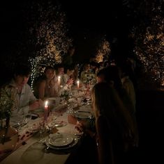 a group of people sitting at a dinner table with candles in the dark, surrounded by trees