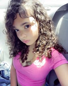 a young woman with curly hair sitting in the back seat of a car, wearing a pink shirt