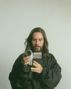 a man with long hair is holding a camera and taking a selfie in front of him