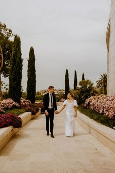 a bride and groom holding hands while walking down a path in front of some flowers