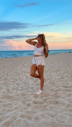 a woman standing on top of a sandy beach next to the ocean with her arms behind her head