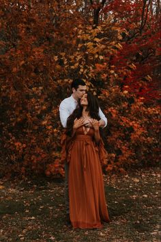 a man and woman standing next to each other in front of trees with orange leaves