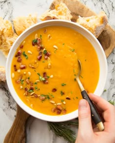 a hand holding a spoon in a bowl of carrot butternut squash soup with toasted bread on the side