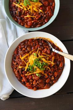 two white bowls filled with chili and cheese on top of a wooden table next to a napkin