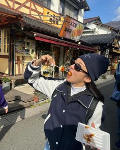 a woman eating food while standing in front of a building on the side of a road