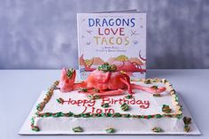 a birthday cake with a dragon on it and a book about love tacos in the background