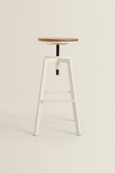 a white stool with a wooden seat and black feet rests against a plain wallpapered background