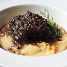 a white bowl filled with mashed potatoes covered in gravy and topped with a sprig of rosemary