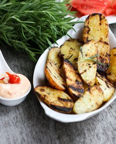 grilled potatoes in a white bowl with dip and fresh herbs on the side for garnish