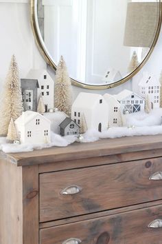 a wooden dresser topped with white houses and snow covered trees in front of a round mirror