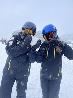 two people standing in the snow with helmets on and one holding something up to their face