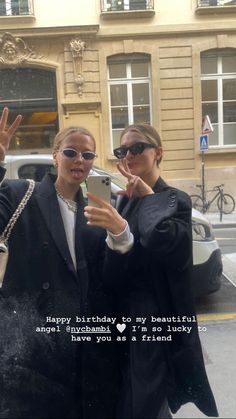 two women standing next to each other in front of a building holding up their cell phones