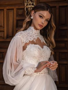 a woman wearing a white wedding dress with sheer sleeves and an open neckline, standing in front of a wooden door