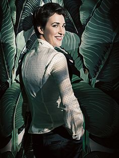 a woman standing in front of a green plant with leaves on the wall behind her