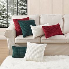 a white couch with red and green pillows in front of a snowy tree outside the window