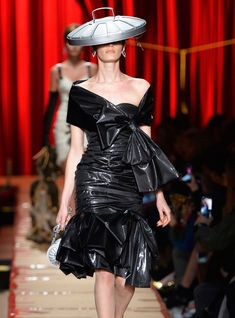 a woman walking down a runway wearing a black dress and hat with large white hats on her head