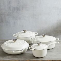 white cast iron cookware set with lids and spoons on a wooden table in front of a concrete wall