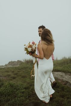 a man and woman walking in the grass with flowers on their back, holding each other