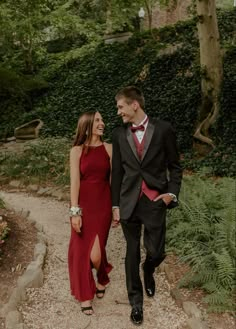 a man and woman in formal wear walking down a path through the woods holding hands