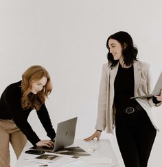 two women standing around a table with laptops and papers in front of each other