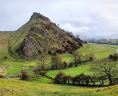 a lush green hillside covered in trees next to a winding road on a cloudy day