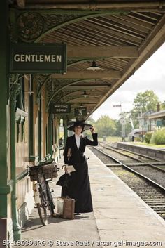 a woman in a long black coat and hat stands on the train platform with her luggage