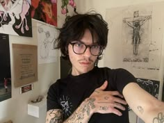 a man with tattoos and glasses standing in front of a wall full of pictures on it