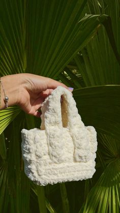 a hand holding a white crocheted purse in front of a palm leaf background