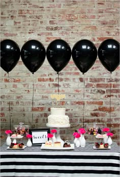 a table topped with black balloons next to a cake and cupcakes in front of a brick wall