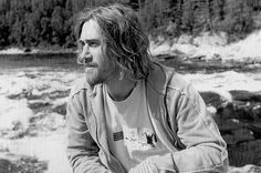 a man with long hair sitting next to a river