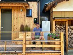 a man sitting on a wooden bench in front of a small house with wood sidings