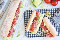 two sub sandwiches sitting on top of a table next to tomatoes and lettuce