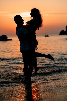 a man carrying a woman on his back while standing in the water at sunset by the beach