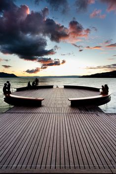 people are sitting on the edge of a wooden pier at sunset or dawn with clouds in the sky