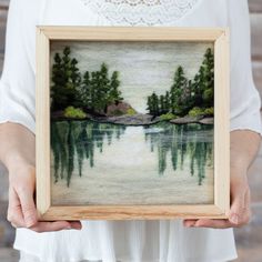 a woman holding up a painting with trees in the water and rocks on the shore