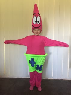 a little boy in a pink costume with green pants and a red hat standing next to a white wall