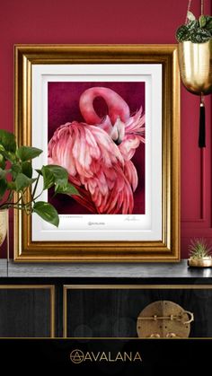 a pink flamingo in a gold frame sitting on top of a table next to a potted plant