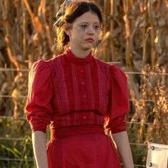 a woman in a red dress standing next to a fence and cornfield behind her