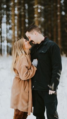 a man and woman standing in the snow with trees behind them, one is kissing the other