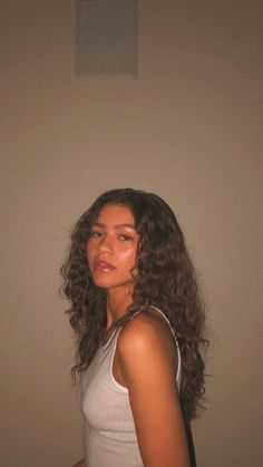 a woman with long curly hair standing in front of a wall and looking off to the side