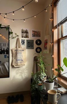 a room with some plants and pictures on the wall next to a window that has lights strung from it