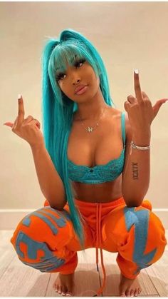 a woman with blue hair and tattoos sitting on the floor wearing an orange outfit holding her fingers up