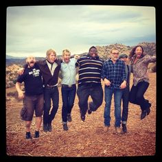 a group of people jumping in the air on top of a dirt field with mountains in the background