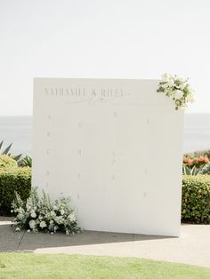 a white sign with flowers on it sitting in front of some bushes and grass next to the ocean