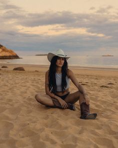 a woman sitting on top of a sandy beach next to the ocean wearing a cowboy hat