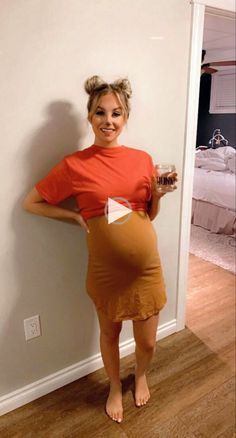 a woman in an orange shirt and brown skirt standing next to a wall holding a bottle